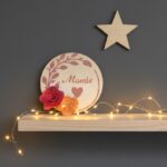 Magnet rond "MAMIE" avec fleurs rose et saumon sur une étagère en bois lumineuse.